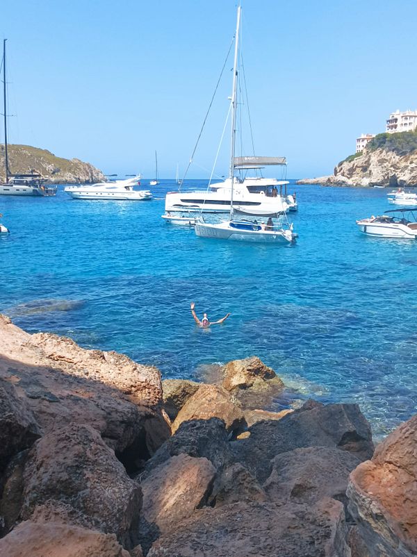 A person is snorkeling in a clear blue bay surrounded by boats and rocky shores.