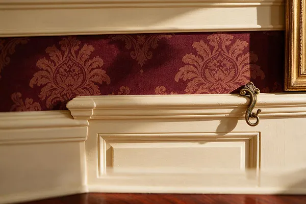 A close-up of a traditional interior detail featuring cream painted wainscoting and trim beneath burgundy damask wallpaper. A small brass hook and the edge of a gilt frame add ornate accents, while warm light highlights the layered moldings and classic finish.