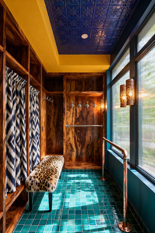 A vibrant mudroom with ribbed-glass windows and teal ceramic tile flooring. Richly grained wood lockers feature chrome hooks and navy-and-cream ikat curtains, with a leopard-print bench below. A mustard soffit frames a glossy cobalt pressed-tin ceiling, while patterned sconces and a patinated copper handrail add glow and texture, creating a layered, maximalist look.