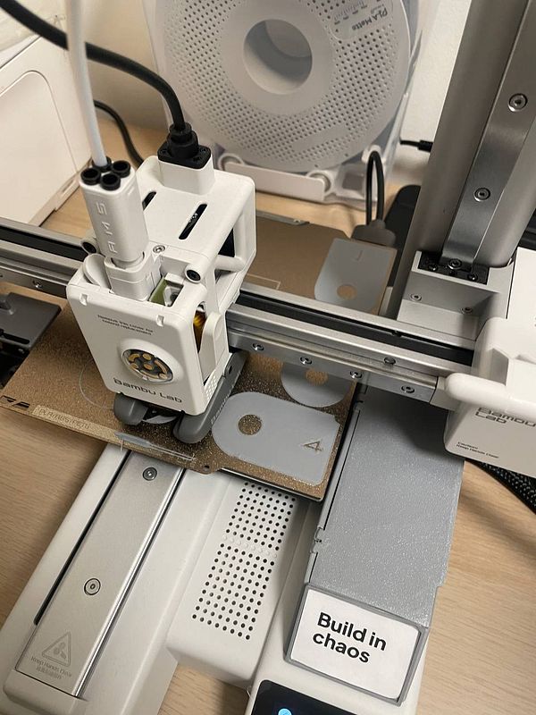 A photograph of a 3D printer in operation with partially printed objects on the build plate and a sticky note attached.