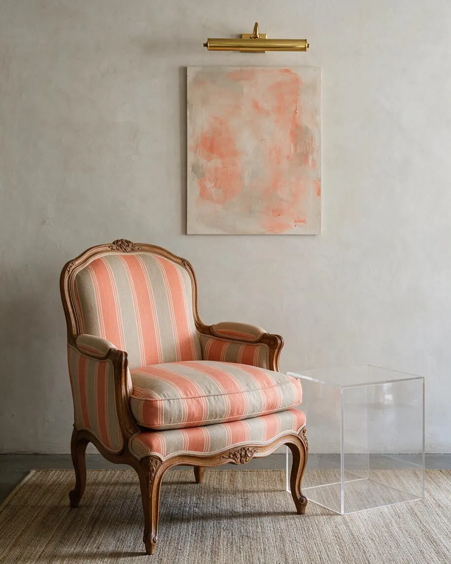 A vintage upholstered bergère chair with carved wooden frame and coral-and-cream striped fabric sits on a neutral woven rug. Above it hangs a small abstract blush painting beneath a brass picture light, while a clear acrylic side table adds a modern contrast.