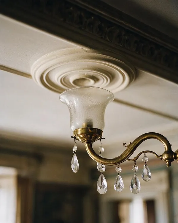 A close-up of a Victorian-style wall sconce with a curved brass arm, frosted glass shade, and dangling crystal pendants. It is mounted beneath an ornate ceiling medallion, with a softly blurred interior background.