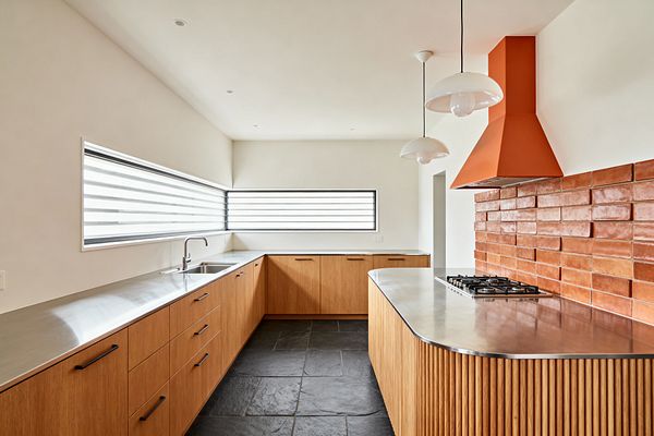 A Mid-Century Modern kitchen with a U-shaped layout features warm wood cabinetry with black pulls, a curved peninsula clad in fluted wood, and seamless stainless-steel countertops with an integrated sink and gas cooktop. A glossy terracotta brick backsplash and matching orange range hood add color against warm white walls, while slate floors and long horizontal corner windows with striped shades and two white globe pendants create a bright, clean look.
