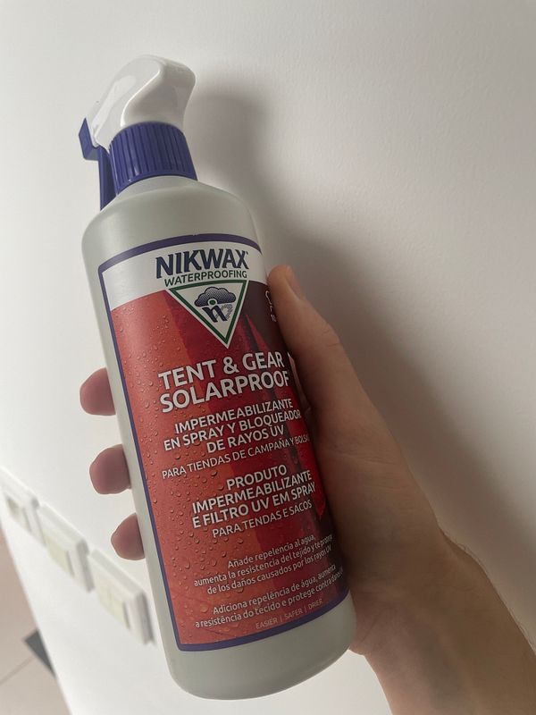 A hand holds a bottle of Nikwax Tent & Gear Solarproof spray against a plain wall.