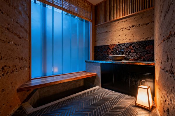 A Japanese-inspired kitchen nook features rammed-earth walls, a patinated copper counter with a round stone vessel sink, and black cabinetry set beneath a backsplash of textured volcanic rock. Soft blue light filters through frosted vertical glass beside a cedar bench, while bamboo slats and a woven shade add warmth; a glowing paper lantern and charcoal herringbone tile floor create a tranquil, minimalist atmosphere.