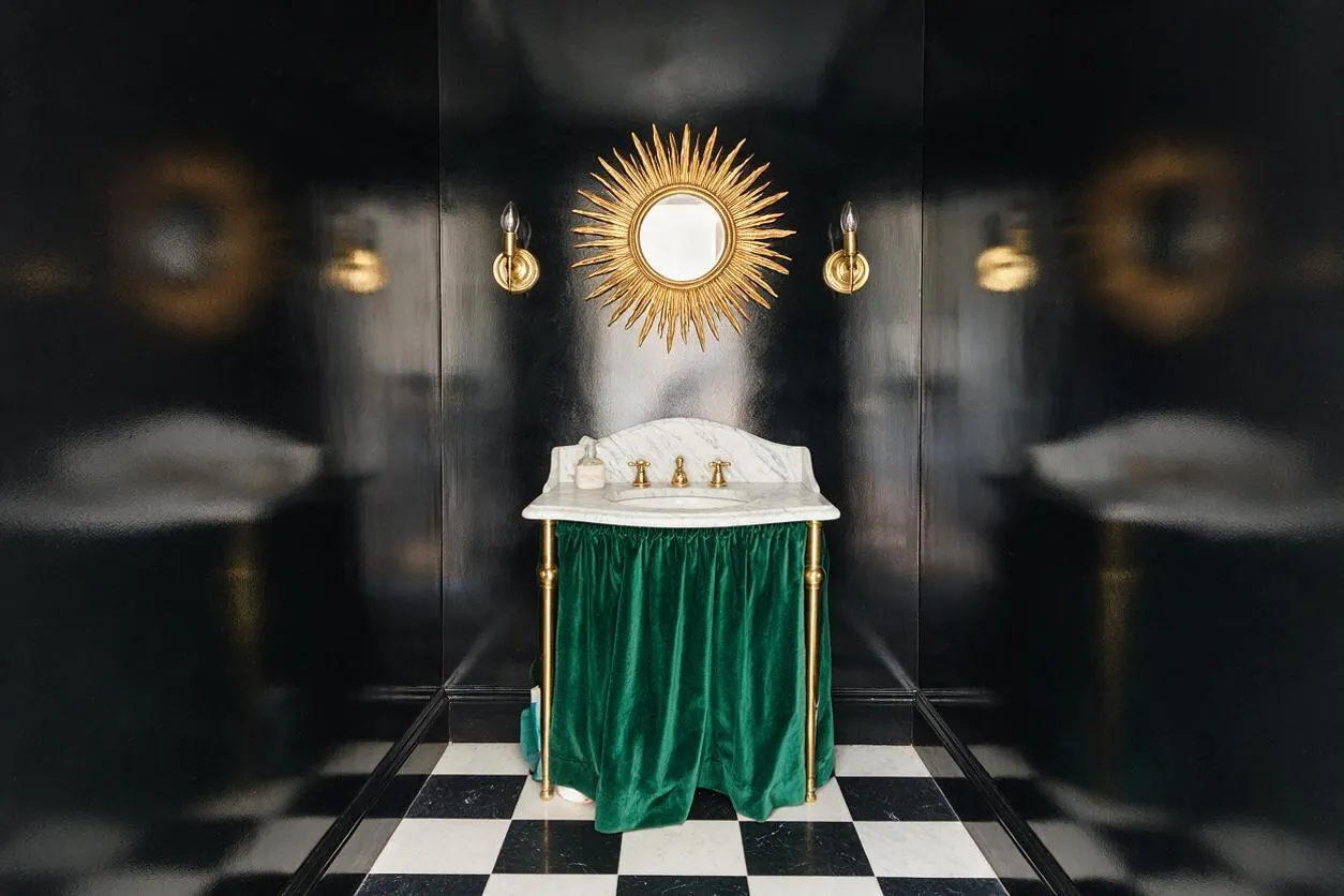 A dramatic powder room with glossy black walls, a black-and-white checkered floor, and a small marble sink wrapped in emerald green velvet. A gold sunburst mirror and matching brass sconces add Hollywood Regency glamour.