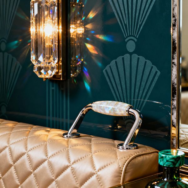 Close-up of a vintage glam home gym detail featuring a champagne-colored, diamond-quilted leather bench with a chrome handle capped in mother-of-pearl. A deep teal, high-gloss panel with Art Deco fan motifs sits behind, while a brass-mounted crystal sconce casts warm light and rainbow refractions across the surface; an emerald, malachite-patterned bottle rests nearby.