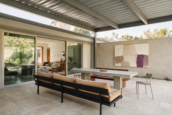 A modern backyard patio with a metal pergola roof, large stone pavers, and a long black-framed outdoor sofa with beige cushions. A concrete dining table with wood bench seating sits beside a minimalist wall-mounted abstract painting, while sliding glass doors open to the interior living room.