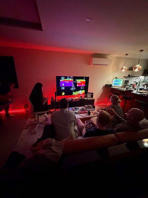 A group of people gathered in a cozy living room, playing a colorful multiplayer video game on a large TV with red ambient lighting.