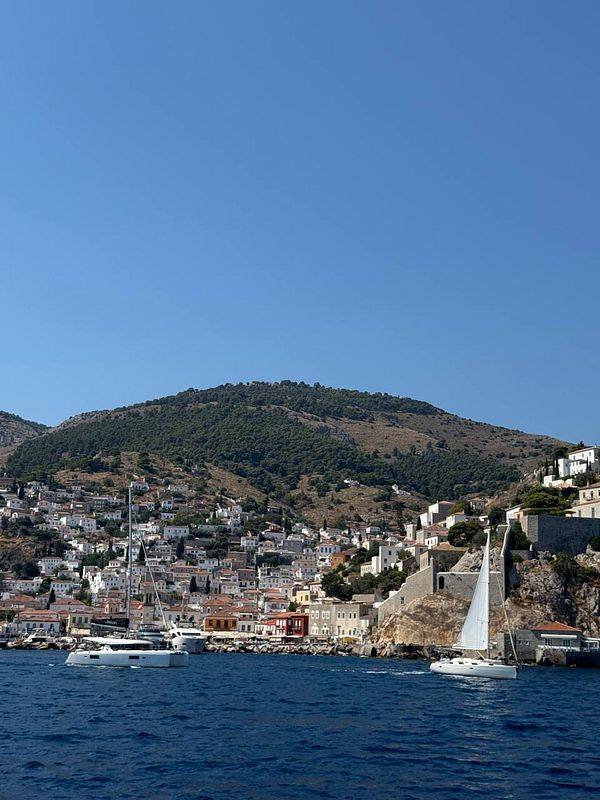 A scenic waterfront view from Hydra island with white buildings and sailboats along the shoreline under a partly cloudy sky.