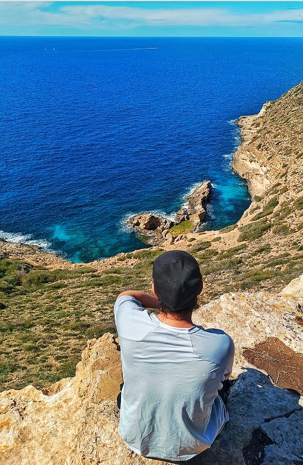 A person sits on a rocky ledge overlooking a vibrant blue sea and rugged coastline.