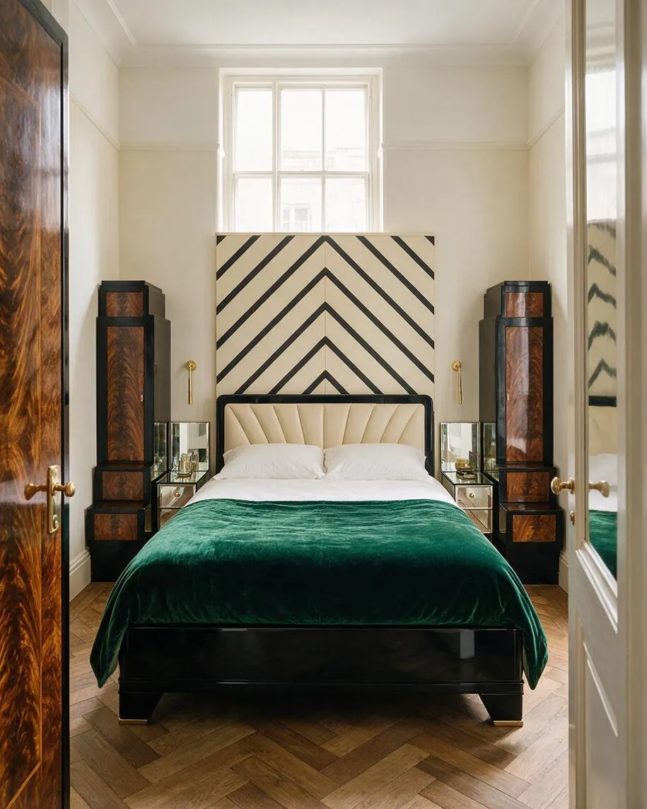 A narrow guest bedroom in Art Deco style featuring a black bed frame, cream upholstered headboard, and a deep green velvet throw. A bold chevron-patterned wall panel rises behind the bed, flanked by mirrored bedside tables and tall lacquered wood cabinets with rich burl wood detailing.
