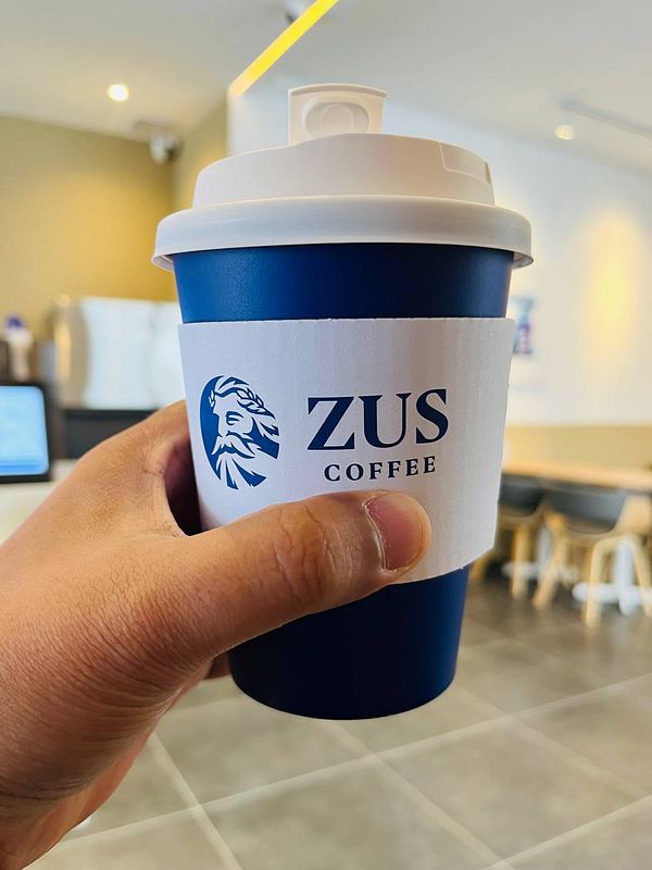 A hand holds a blue coffee cup with a logo in a modern cafe setting.