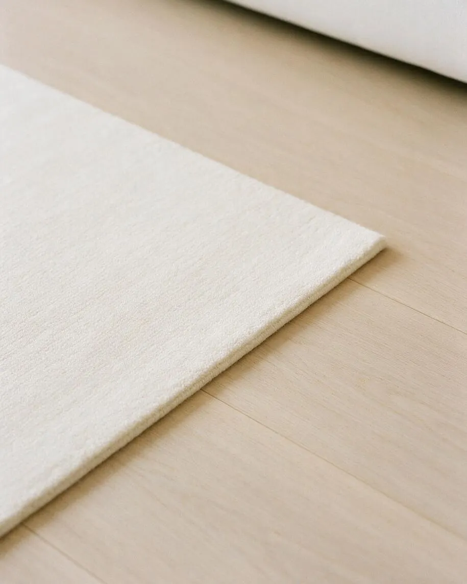 A close-up view of a soft cream rug with a low, plush texture laid over a light natural wood floor. The image highlights the clean edge of the rug, subtle wood grain, and a calm neutral palette in a minimalist interior.