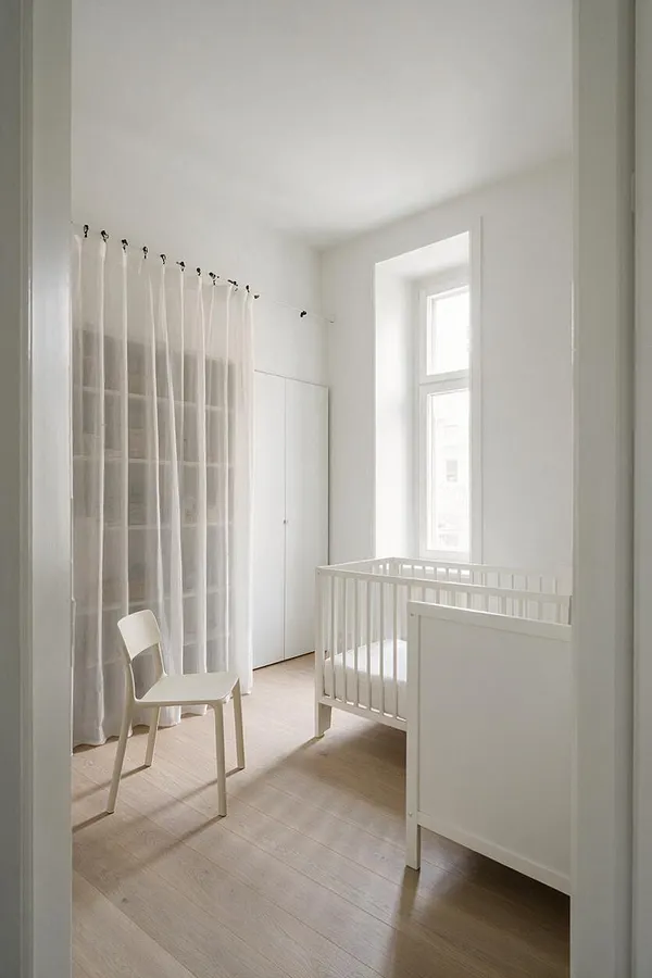 A minimalist nursery with white walls, a white crib beside a tall window, sheer curtains covering a large window, and a small white chair on light wood floors. The room feels airy, sparse, and calm with soft natural light and very few decorative elements.