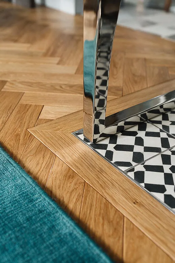 A close-up detail of a warm oak chevron floor surrounding a black-and-white geometric tile inset beneath a polished chrome furniture leg. A teal textured rug borders the wood floor in the foreground, adding color contrast to the eclectic mix of materials and patterns.