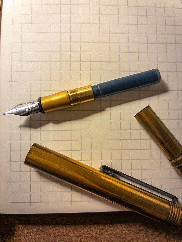 A TRC pen is positioned on a grid-patterned notebook page alongside its cap and another pen.
