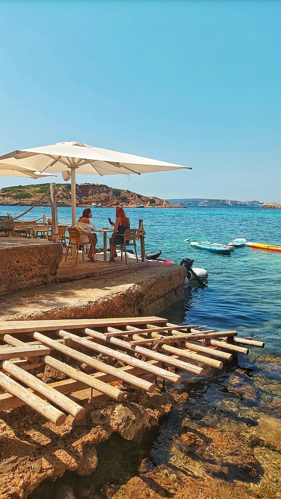 A serene beach scene featuring two women seated at a table under a large umbrella, with a clear blue sea in the background.