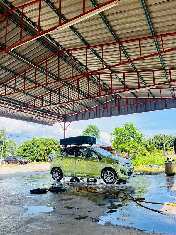 A green car is being washed at a car wash facility under a large roof.