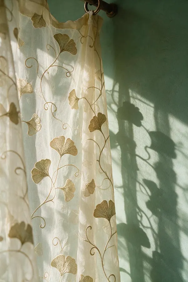 A close-up of translucent cream curtains embroidered with flowing gold ginkgo leaves and curling vine motifs. Sunlight passes through the fabric, casting soft botanical shadows onto a muted green wall, creating a delicate Art Nouveau look.