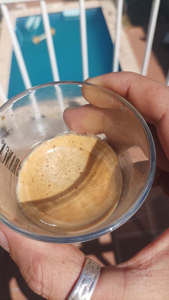 A hand holds a glass of espresso with a swimming pool in the background.