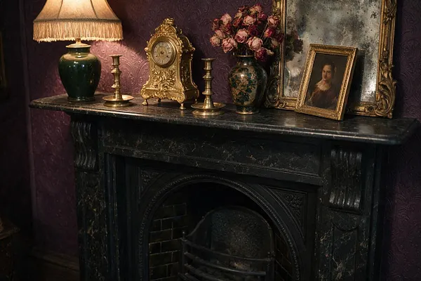 A Victorian-style mantel vignette with a dark marble fireplace surround, topped by a green ceramic lamp with a fringe shade, gilt candlesticks, a gold mantel clock, floral porcelain vase, and framed portrait art. Rich burgundy patterned wallpaper and an ornate gilded mirror create a dramatic, antique atmosphere.