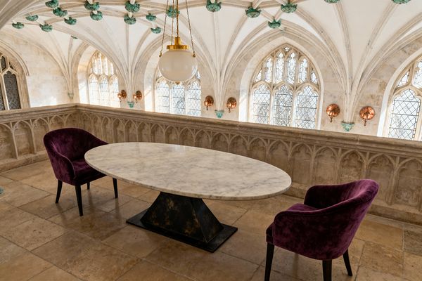 A compact dining vignette set in a Gothic stone gallery with ribbed vaults and pointed arched, leaded-glass windows. An oval white marble table on a blackened pyramidal base is paired with two deep purple velvet armchairs; a brass-chained globe pendant and round copper wall sconces warm the honey-beige limestone floors, carved tracery balustrade, and verdigris ceiling bosses.