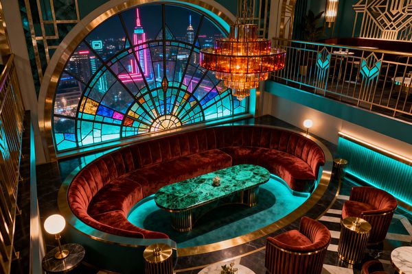 A double-height Art Deco living room features a sweeping curved banquette in oxblood velvet surrounding an oval emerald-green marble coffee table on fluted brass legs, set atop black marble flooring with white geometric inlay. A large arched stained-glass window with fan-pattern panes overlooks a neon-lit cityscape, while teal walls, brass railings, concealed turquoise lighting, an amber tiered chandelier, and small globe table lamps complete the glamorous, jewel-toned scheme.