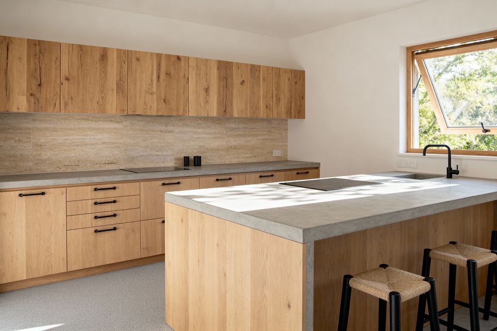 A bright Scandinavian kitchen with flat-front oak cabinets and a matching island topped in light gray concrete. A beige travertine backsplash runs the length of the back wall, matte black pulls and faucet add contrast, and three black stools with woven rush seats line the island beneath a wide wood-framed window bringing in natural light.