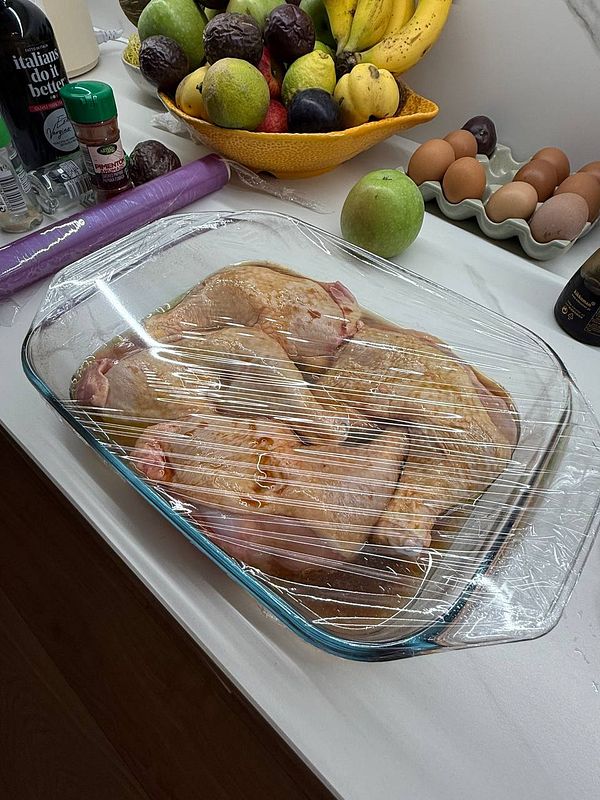 A glass dish containing marinated chicken is covered with plastic wrap on a kitchen countertop.