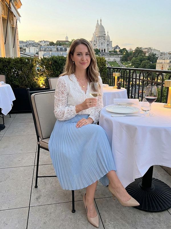 A woman sits elegantly at a restaurant table in Paris, with the Sacré-Cœur Basilica visible in the background.