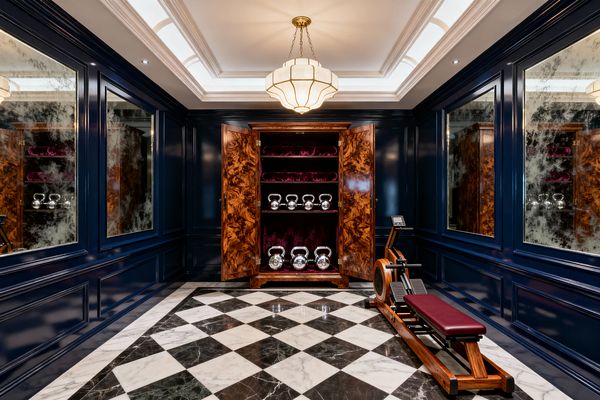 A jewel-box home gym features deep navy lacquered wall paneling inset with large antiqued mirror panels and a central walnut-burl armoire lined in plum velvet holding polished chrome kettlebells. A black-and-white checkerboard marble floor, a solid-wood rowing machine with a burgundy seat, and a tray ceiling with cove lighting and an Art Deco opaline pendant in brass create a richly lit, vintage-glam atmosphere.