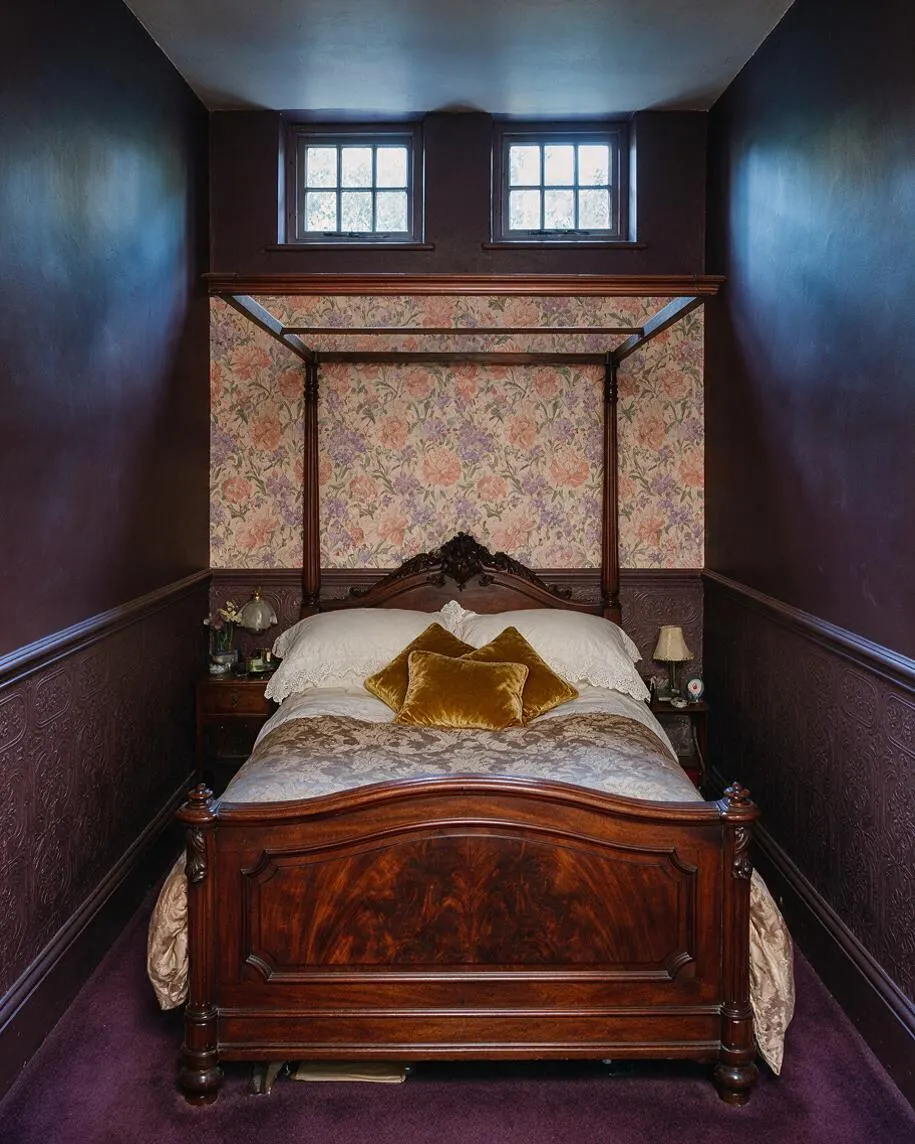 A narrow Victorian-style basement bedroom with dark plum walls, ornate textured wainscoting, and a rich wood canopy bed centered beneath two small windows. Floral wallpaper fills the headboard wall, while white bedding and gold velvet pillows soften the deep, moody palette.