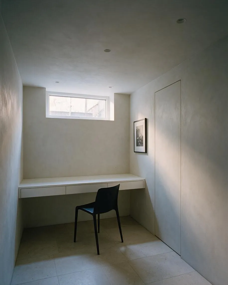 A compact minimalist basement room with smooth white walls, a small horizontal window near the ceiling, and a built-in floating desk with drawers. A single black chair sits beneath the desk, with a framed black-and-white print on the right wall and soft natural light entering the space.