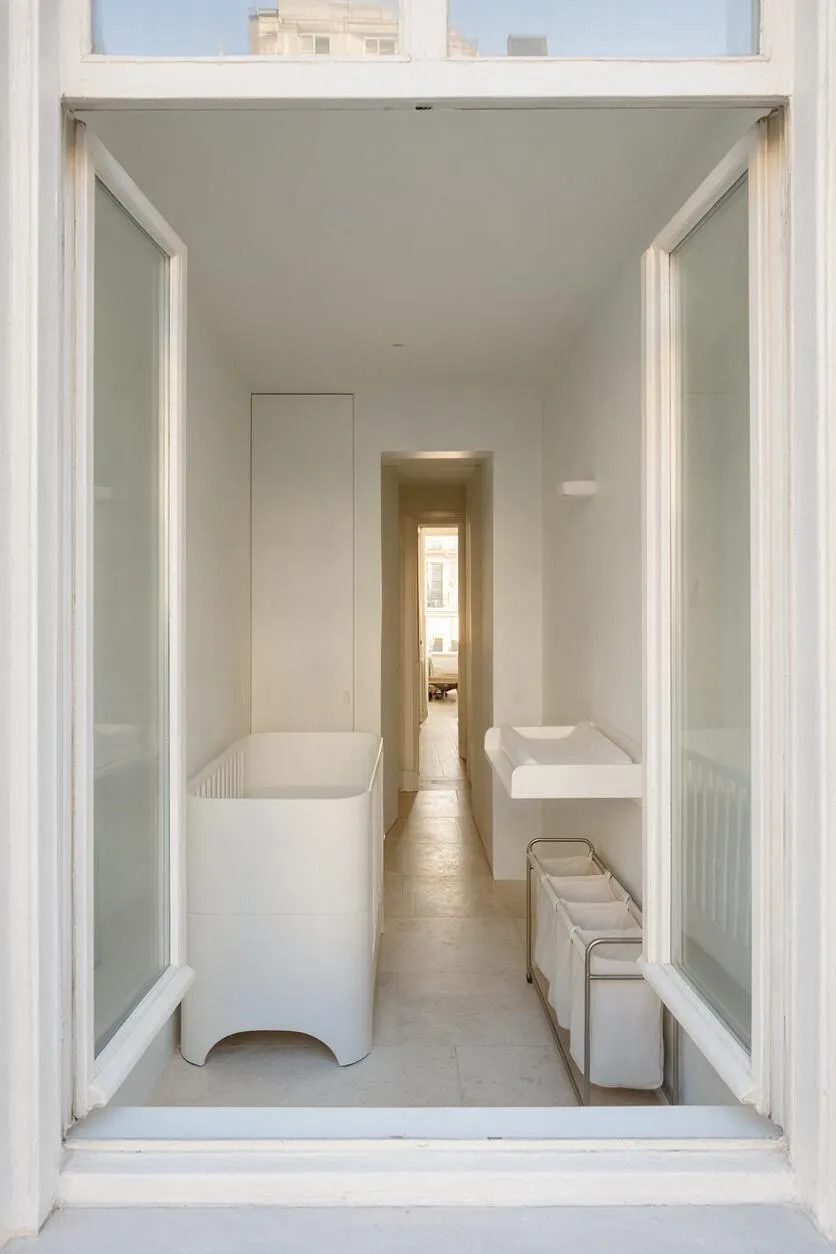 A narrow minimalist nursery seen through open French windows, with white walls, pale stone flooring, and simple baby furniture arranged along both sides. A white crib sits on the left, a changing table on the right, and a slim storage basket below, creating a calm, uncluttered layout filled with soft natural light.