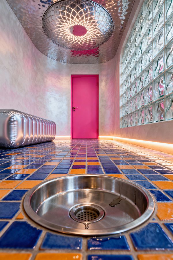A narrow mudroom with a glossy blue-and-orange tiled floor features a circular stainless floor drain in the foreground. A vivid pink door anchors the far wall; to the right runs a wavy glass‑block wall with warm LED base lighting, while a silver inflatable-looking bench sits to the left. Overhead, a mirrored metal ceiling and a domed light cast a kaleidoscopic pattern across softly mottled pink-gray plaster walls, creating a psychedelic atmosphere.