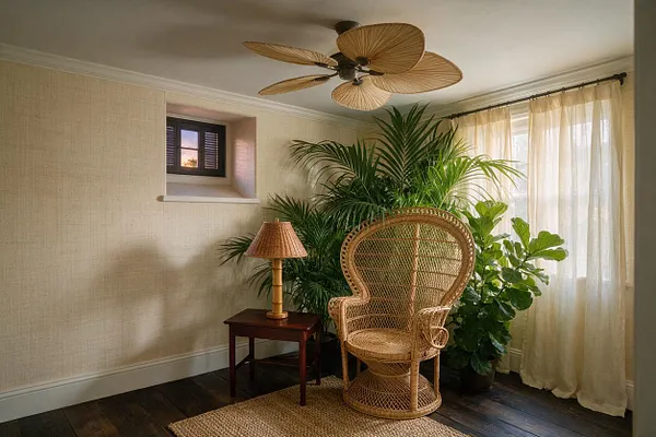 A cozy tropical-style basement corner features a woven rattan peacock chair, a small dark wood side table with a wicker lamp, and several large potted tropical plants grouped near sheer cream curtains. Textured beige walls, a small basement window, dark wood flooring, a jute rug, and a ceiling fan with palm-leaf blades create a warm, airy retreat.