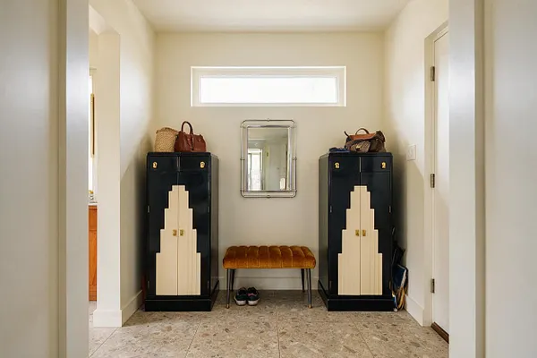 A narrow entryway with cream walls and a high transom window centered above a small mirrored wall piece. Two black cabinets with stepped cream Art Deco inlays flank a rust-gold tufted bench, while terrazzo-style flooring and a few bags and shoes complete the functional space.