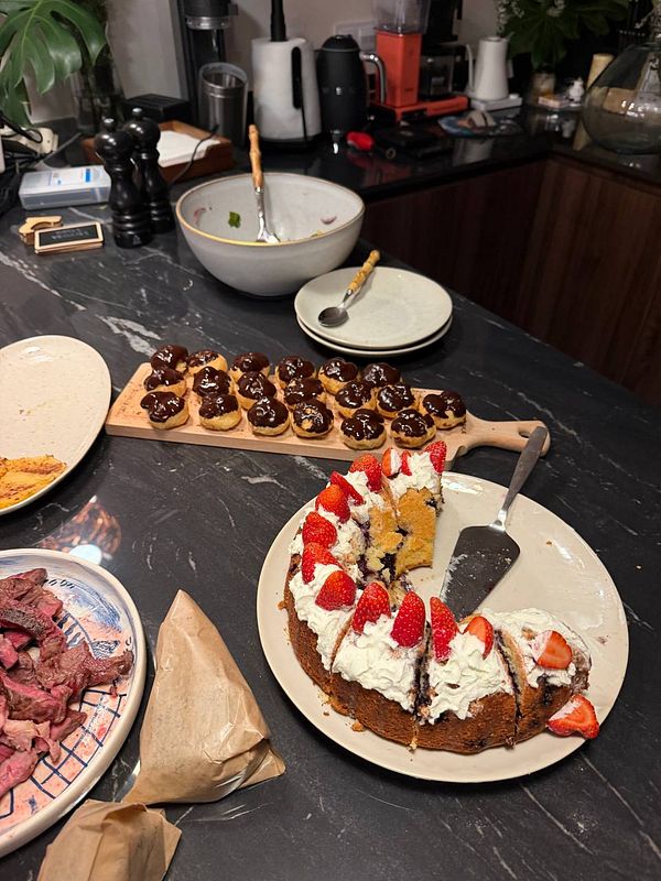 A kitchen counter set with various foods including a sliced strawberry cake, small chocolate-topped pastries, a bowl with utensils, plates, and sliced cooked meat on a plate.