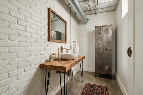 An industrial powder room with a white painted brick wall, a live-edge wood vanity, a white vessel sink, and brass faucet. A weathered metal locker stands at the far end, while exposed ductwork and a concrete floor reinforce the utilitarian loft style.