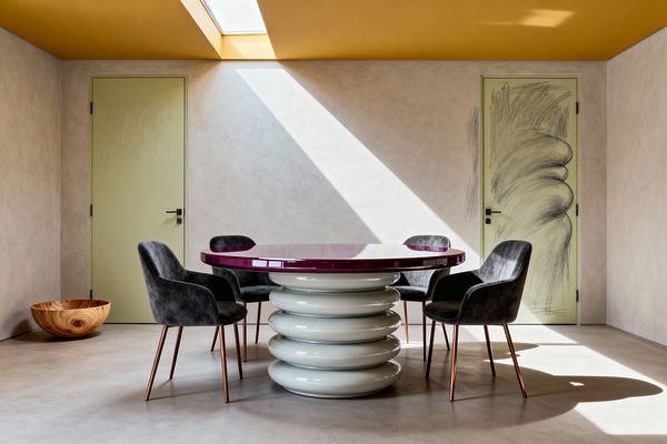 A minimalist dining room features a round table with a glossy plum top and a sculptural base of stacked pale-gray rings, surrounded by four charcoal velvet chairs on copper legs. Creamy plaster walls and a polished concrete floor sit beneath a mustard-yellow ceiling with a skylight sending a sharp diagonal beam of sunlight across the room. Two sage green doors, one overlaid with a loose charcoal sketch, and a wooden bowl accent the scene.