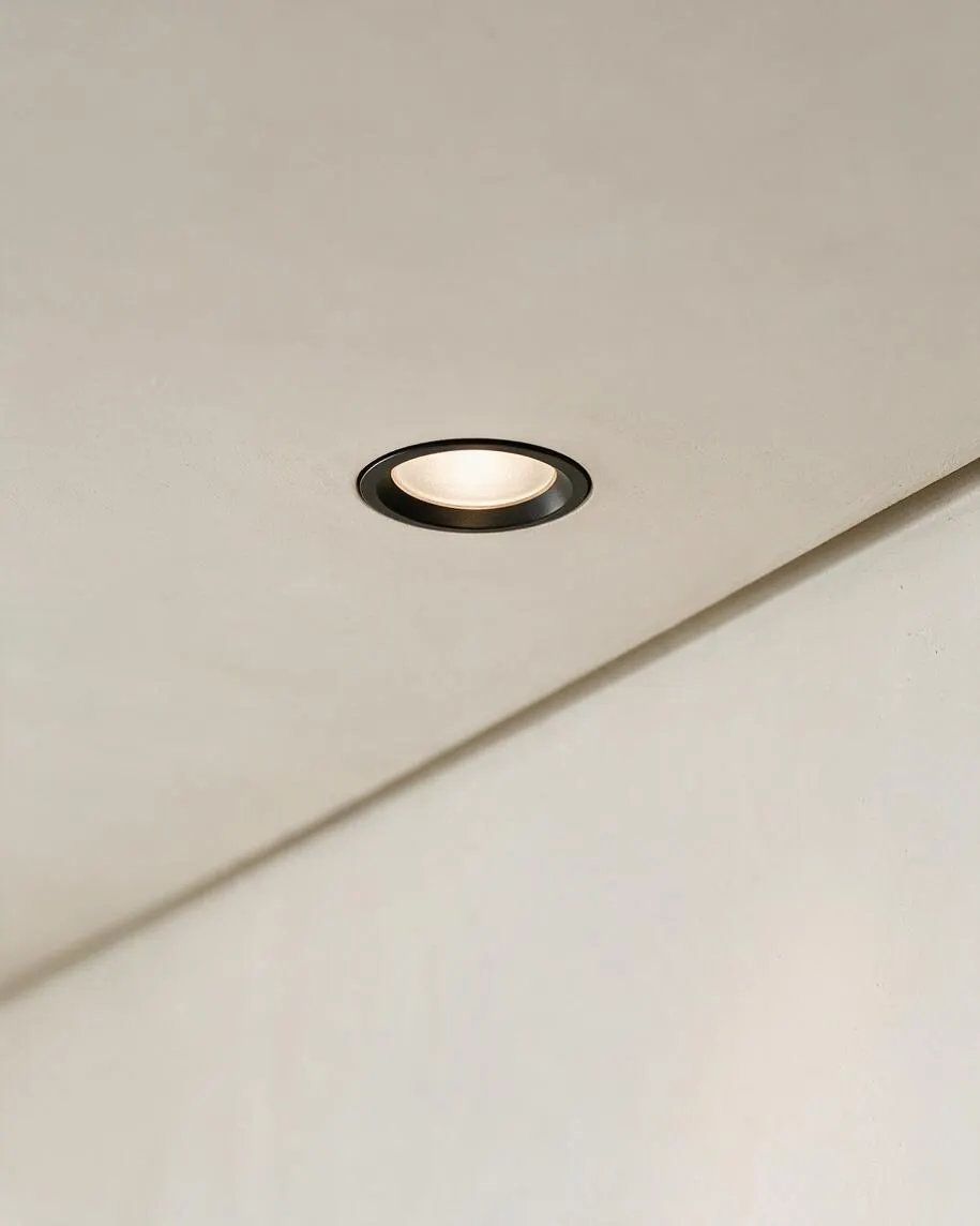A close-up view of a smooth off-white ceiling with a small recessed downlight featuring a dark trim ring and soft warm illumination. A diagonal wall or beam line crosses the lower half of the image, emphasizing the clean, minimal architectural detail.