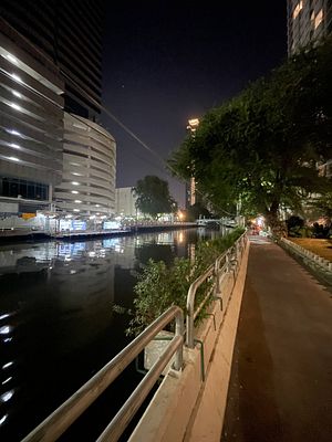 A nighttime riverside walkway with reflections on the water and urban buildings in the background.