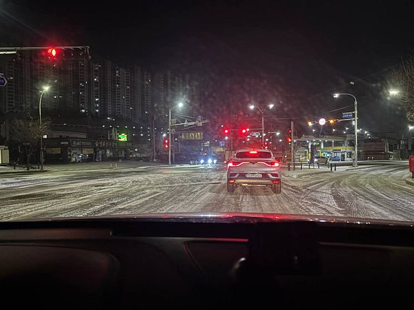 A snowy residential street with a car driving on it and houses on both sides covered in snow.