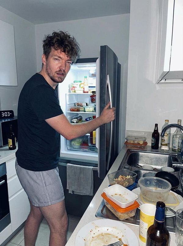 A man in a kitchen is opening a refrigerator while wearing only striped boxers.