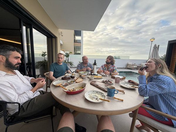 Five people sitting around a circular table on a patio next to a pool, sharing a meal in an outdoor setting during early evening.