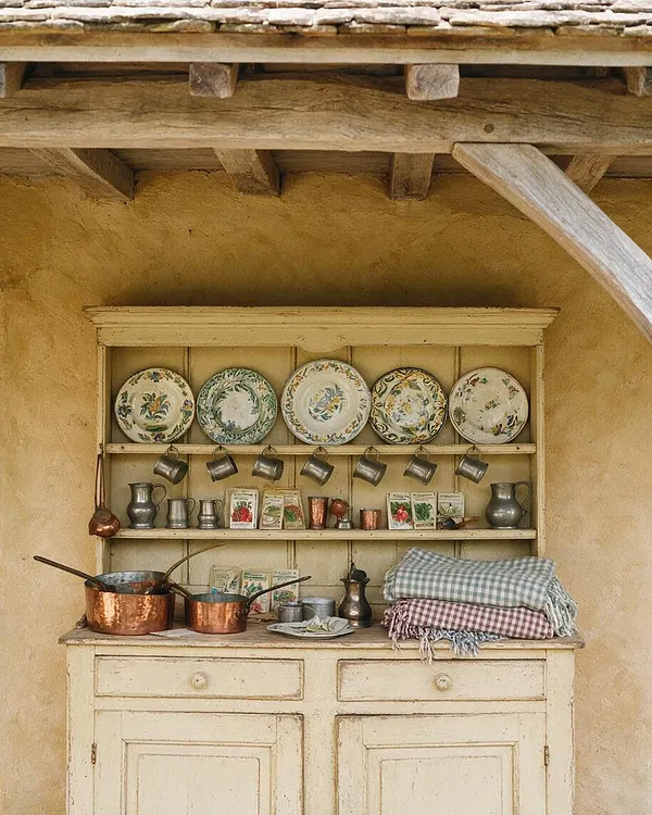 A French Country front porch with a weathered cream hutch set against a warm stucco wall, decorated with floral plates, hanging metal cups, copper cookware, pitchers, and folded gingham textiles. Exposed timber beams and a stone roof edge frame the cozy, antique-inspired arrangement.
