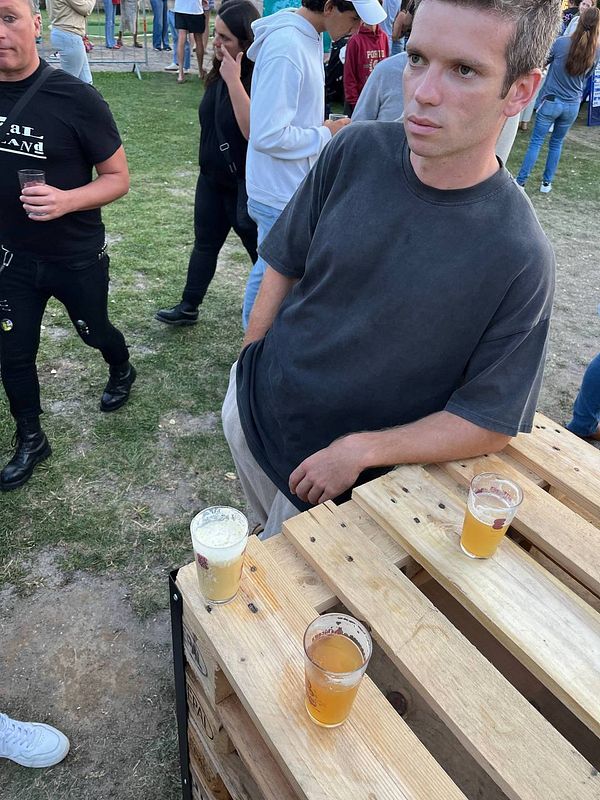 A young man stands next to a wooden table with three cups of beer at a festival.