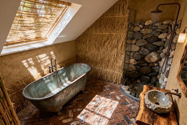 A rustic attic bathroom with a sloped ceiling and skylight filtered by a bamboo blind casts dappled light over a verdigris metal freestanding tub on a herringbone brick floor. At right, a river-stone shower with exposed patinated pipe and rainfall head sits beside a live-edge wood vanity with a rough stone vessel sink, while clay-textured walls and a straw-clad partition emphasize natural materials and earthy tones.