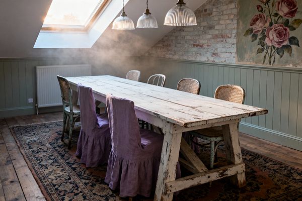 A shabby chic dining room features a long, distressed white wooden trestle table on a faded patterned rug and wide-plank floors. Mismatched seating includes cane bistro chairs and painted metal chairs, some covered with dusty lavender linen slipcovers. Sage green beadboard wainscoting runs beneath a limewashed brick wall and a vintage rose mural. Three ribbed glass pendant lights hang above, while a large roof skylight washes the space with soft natural light.